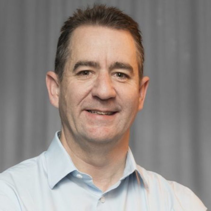A person with short hair wearing a light-colored shirt stands against a plain backdrop, looking at the camera with a slight smile.