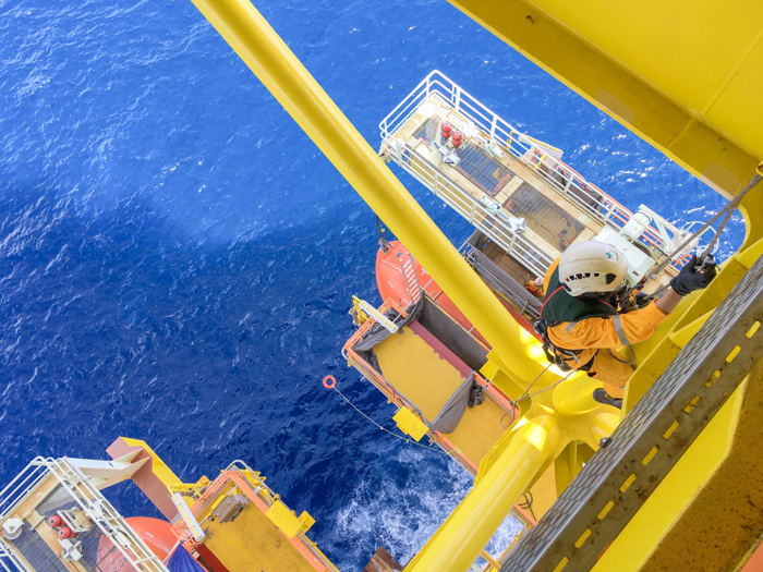 Worker in safety gear climbing yellow structure on an offshore oil platform, with deep blue ocean in the background.