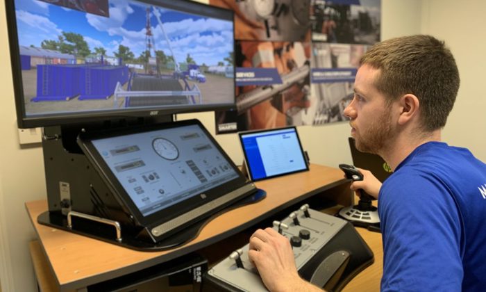 A person operates a control simulator with multiple screens displaying graphics and controls, simulating an industrial environment, in a training room with posters on the wall.