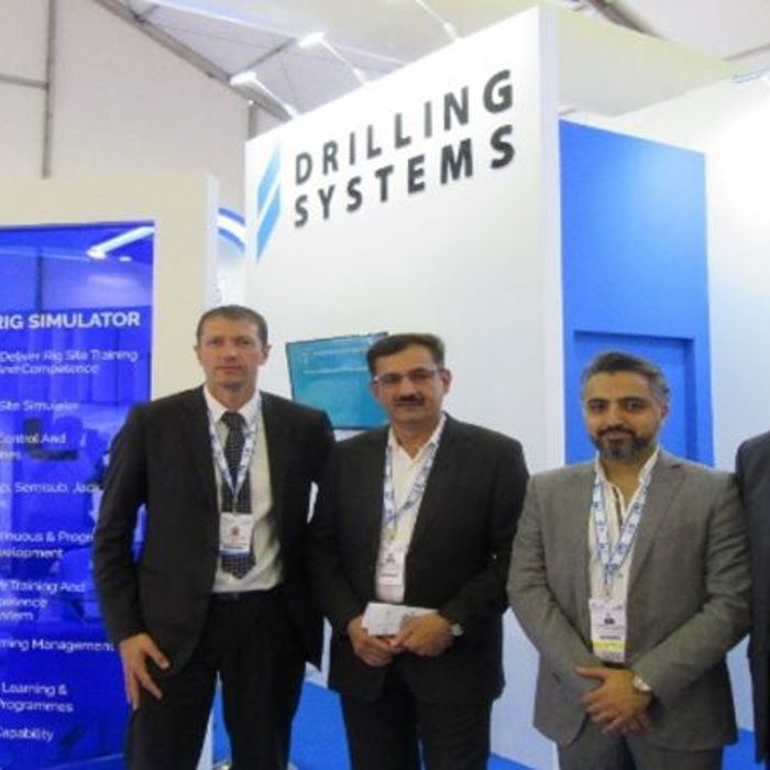 Four men in business attire stand in front of a Drilling Systems booth at a trade show.