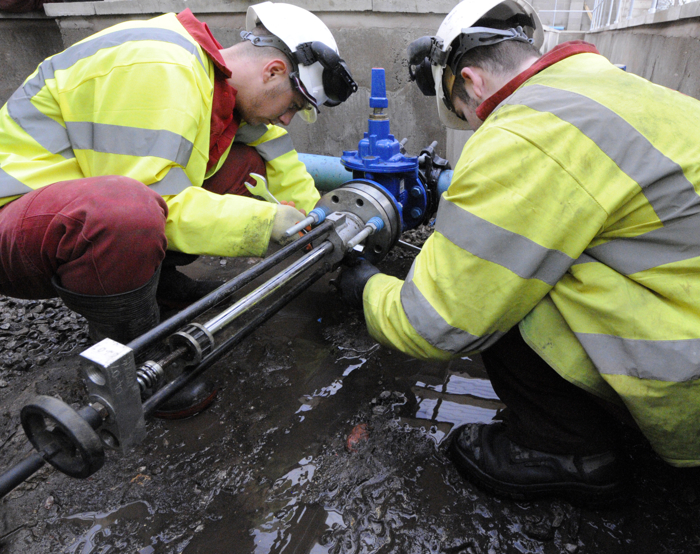 Two workers in high-visibility jackets and helmets are training while inspecting or repairing an industrial pipe outdoors, surrounded by muddy ground.