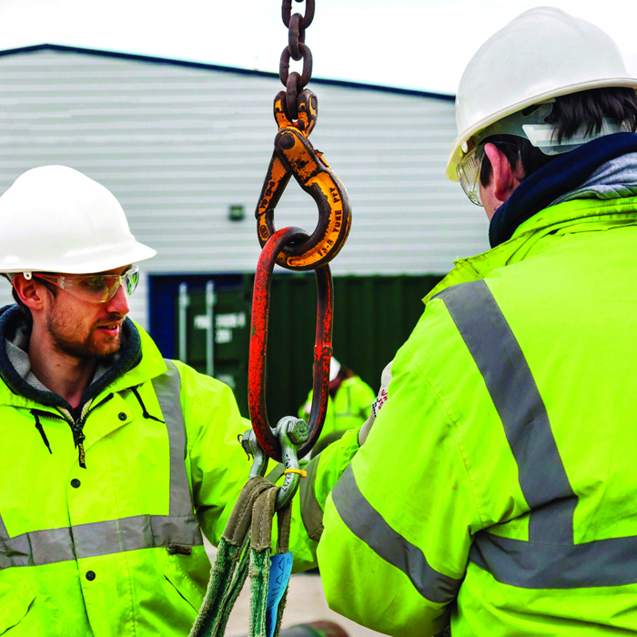 Two construction workers in high-visibility jackets and helmets are skillfully handling a large hook and chain outdoors near a building, showcasing their training.