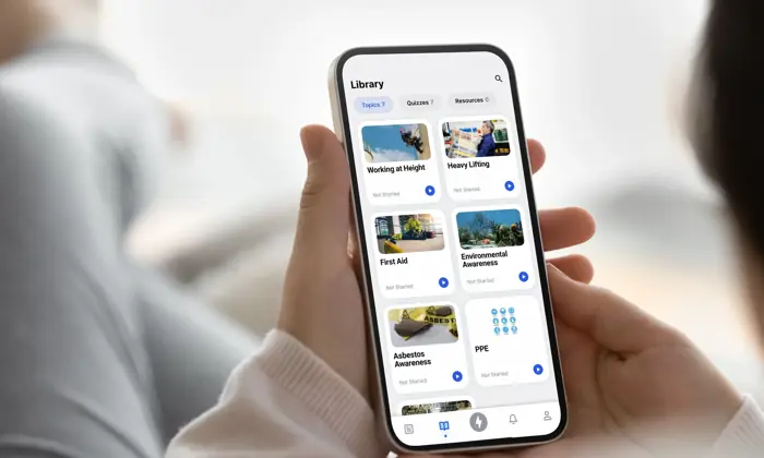 Person using a smartphone app to browse a library of courses including "Working at Height," "First Aid," "Asbestos Awareness," among others, with options categorized under various topics and sectors.