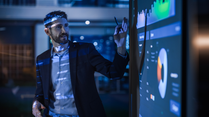 A man in a business suit points at a large interactive screen displaying graphs and charts in a modern office setting.