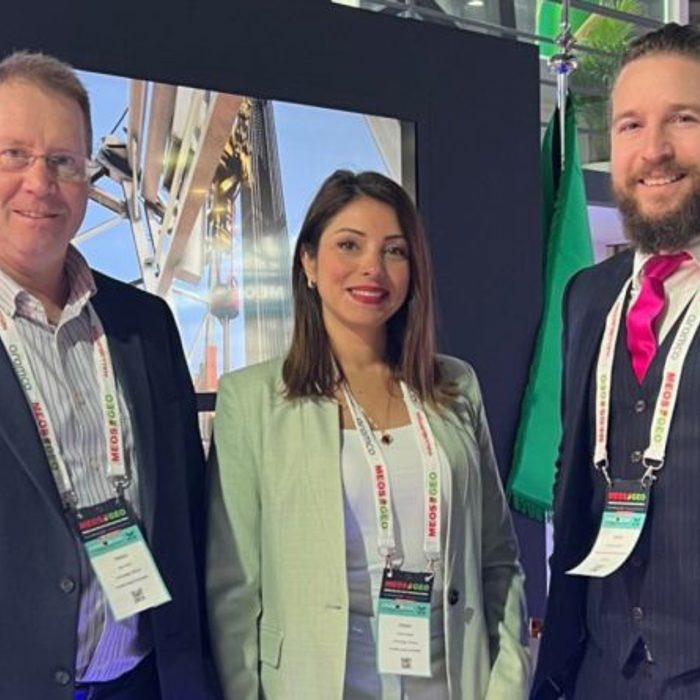 Three people dressed in business attire stand together, each wearing conference badges around their necks. A screen displaying an outdoor scene is visible in the background.