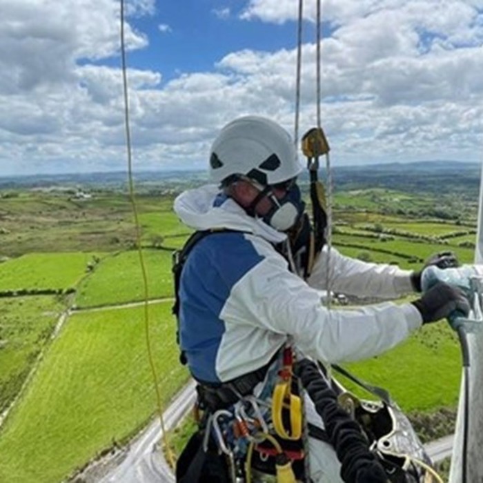 A person wearing safety gear and a helmet works on a structure high above a rural landscape with green fields under a partly cloudy sky.