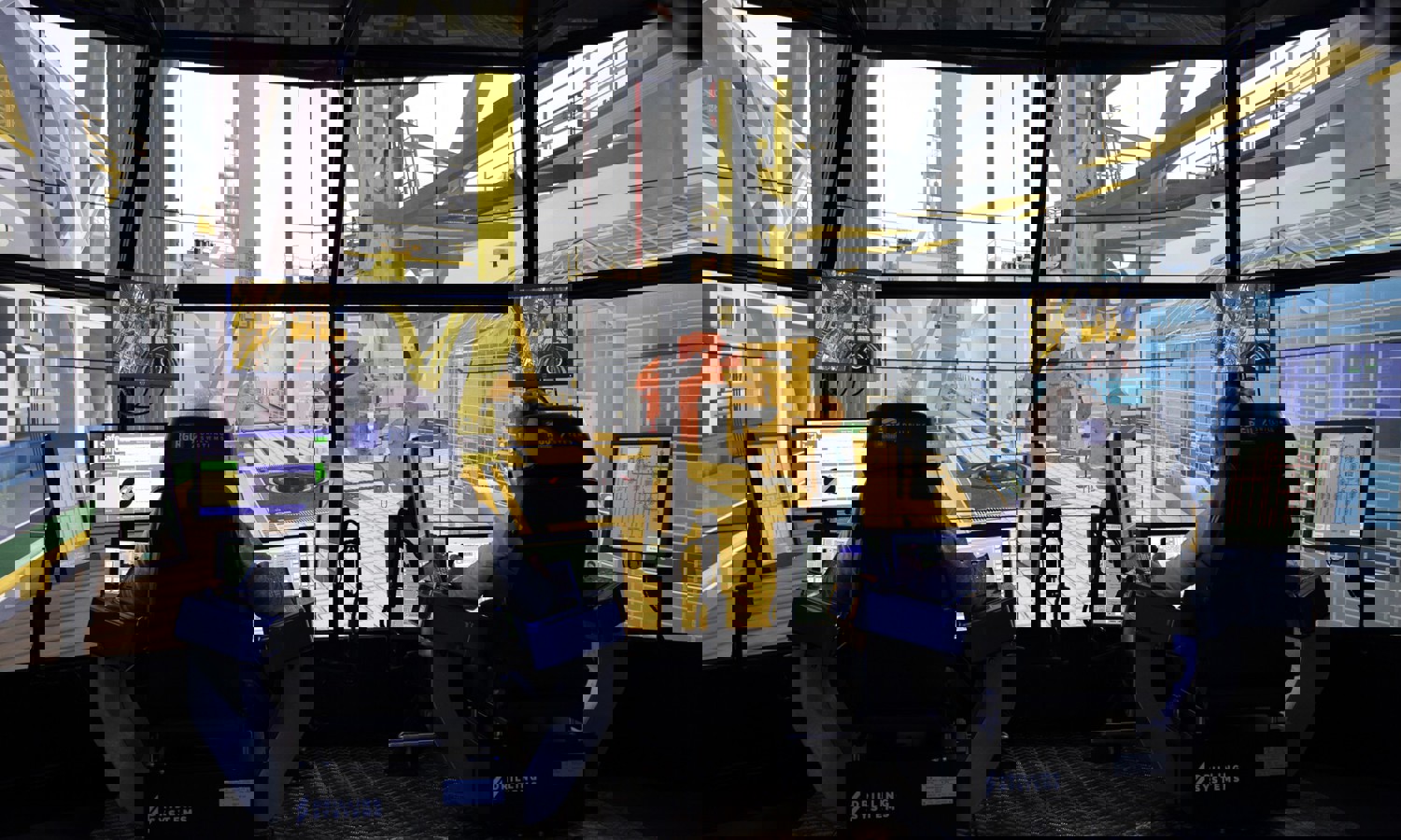 Two people sit at control stations with multiple monitors, operating a high-tech drilling simulator. Large screens simulate an offshore drilling environment around them.