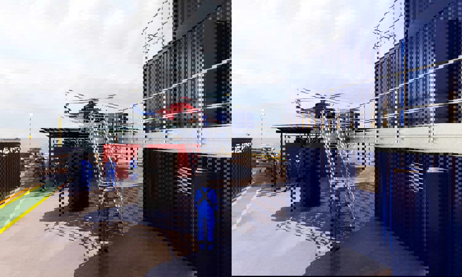 Helicóptero numa plataforma com trabalhadores de uniforme azul nas proximidades, situado ao lado de contentores de metal.