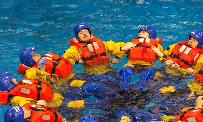 People in yellow suits and helmets float in a pool in a circular formation, wearing orange life vests as part of their training. A person in black stands nearby.