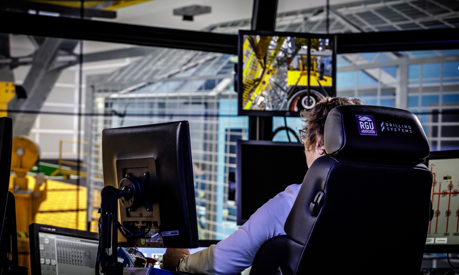 A person operates heavy machinery using a control panel and monitors at an industrial facility.