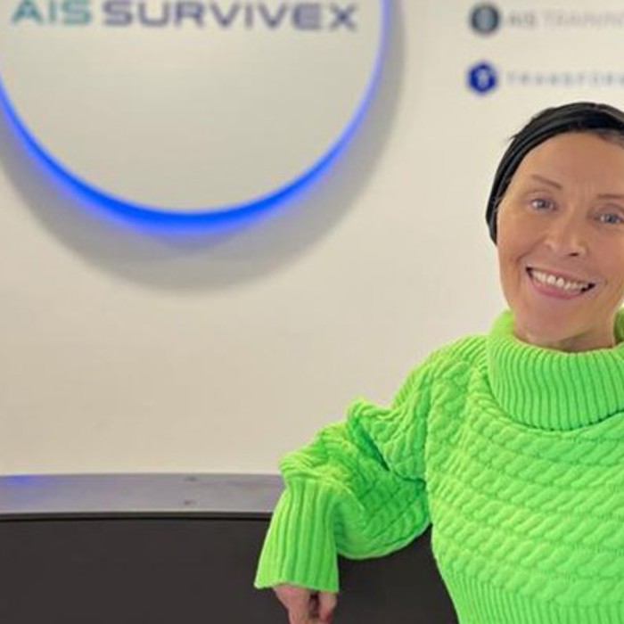 A person with short hair, wearing a bright green sweater, stands smiling near a counter, with a logo reading "AIS Survivex" on the wall behind them.