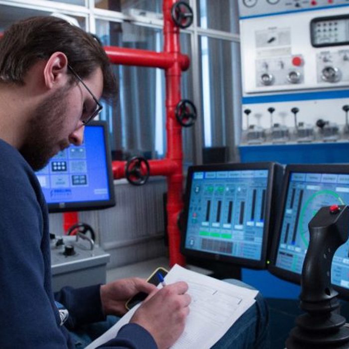 Person in a blue shirt taking notes on paper, surrounded by multiple computer screens displaying graphs and gauges in what appears to be a control room.