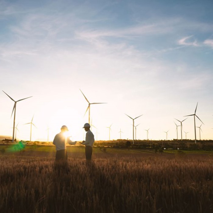 Two people standing in a field at sunset with several wind turbines in the background.