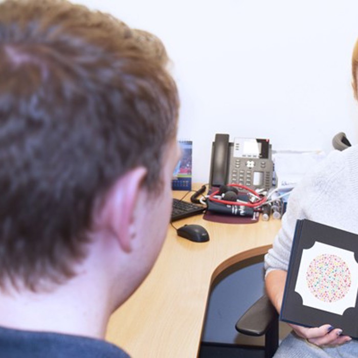 A healthcare professional holds and discusses an Ishihara color blindness test booklet with a patient in an office setting.
