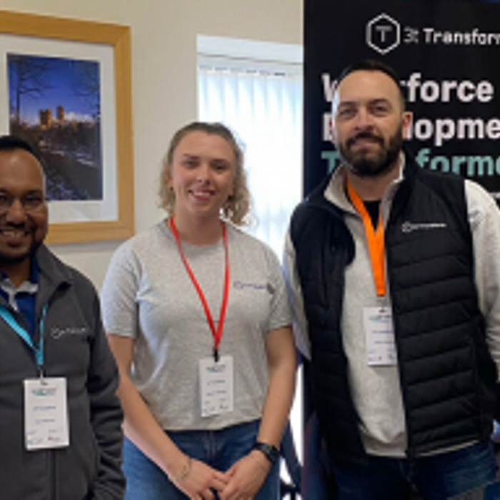 Three people wearing name badges stand together indoors, smiling at the camera. Behind them is a banner that partially reads, "Workforce Development Transformed.