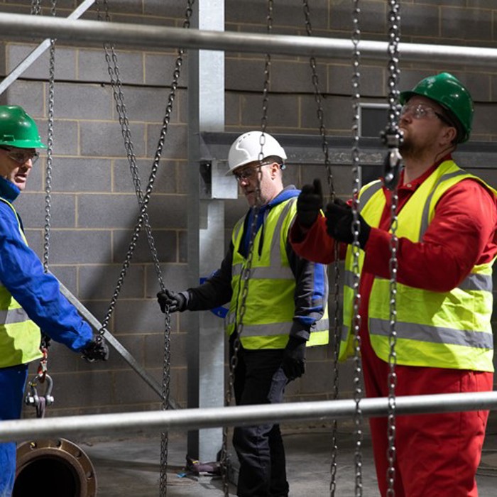 Three workers in high-visibility vests and hard hats operate chains and pulleys on a construction site.