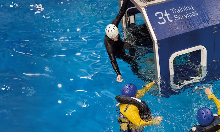 Two individuals in yellow waterproof outfits and helmets receive assistance from a person in a wetsuit and white helmet, emerging from a blue training module labeled "3t Training Services" in a pool.