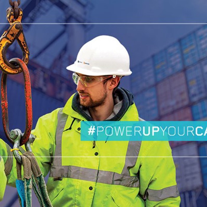 Two workers with safety gear and helmets handling a large hook in front of stacked shipping containers. Text overlay reads: "#POWERUPYOURCAREER".