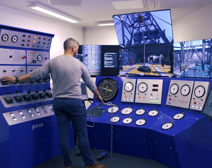 A person operates a blue industrial control panel with multiple screens and gauges, likely in a simulation or control room environment.