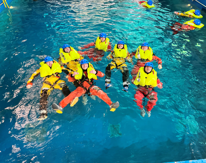 Seven individuals in bright yellow flotation suits and blue helmets float in a swimming pool, practicing water safety or rescue training techniques as precise as those performed by the military.