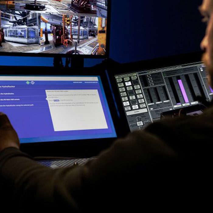 A person operates multiple screens in a control room. One screen displays control options, while another shows surveillance footage of an industrial facility.