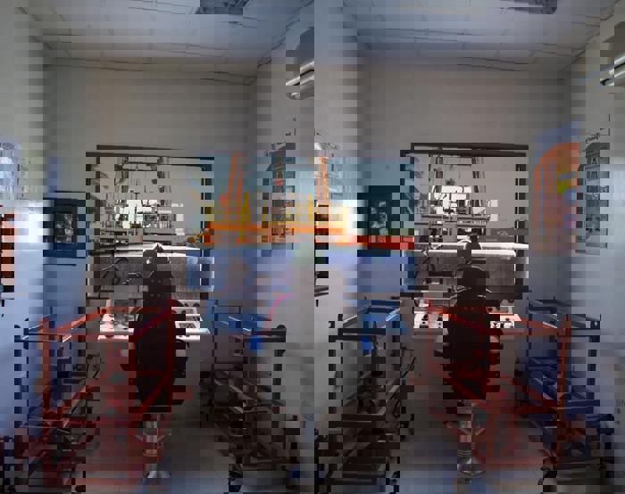 Interior of a control room with a person seated at a console, monitoring a crane lifting a container outside. Red equipment frames flank the room, and informational posters are on the walls.