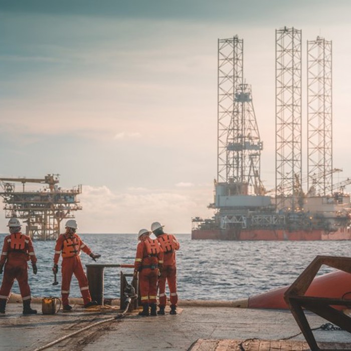 Four workers in orange suits stand on an offshore platform with oil rigs visible in the background under a cloudy sky.