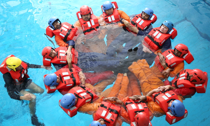 A group of people wearing life vests and helmets form a circle while floating in a pool during a water safety training exercise.