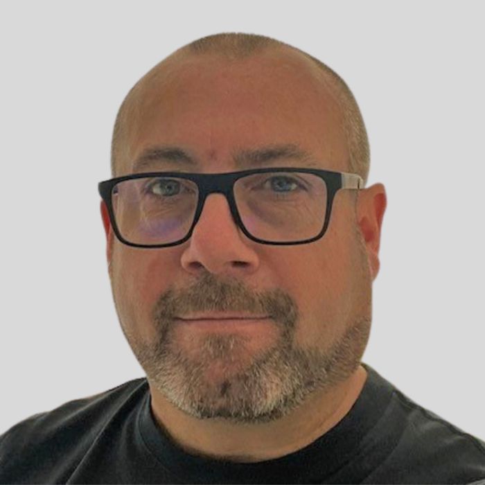 A bald man with glasses, a short beard, and a serious expression is looking at the camera. He is wearing a dark shirt and the background is plain white.