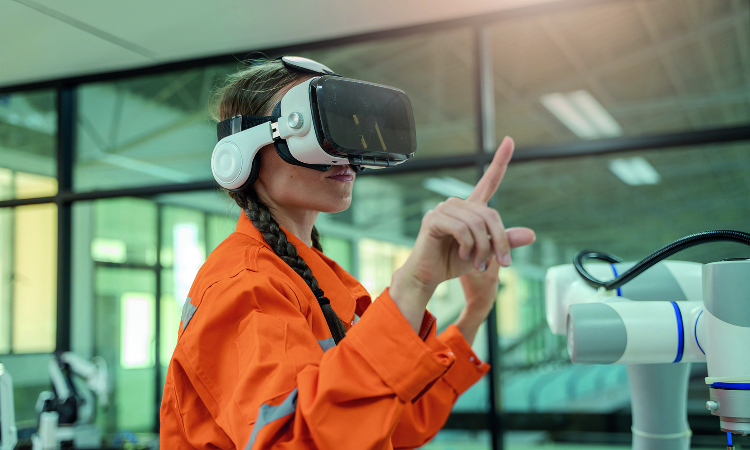 A person wearing an orange jacket and VR headset interacts with a robotic device in a modern industrial setting.