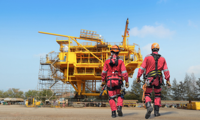 Two workers in red protective gear walk towards a large yellow oil rig under a clear blue sky, exemplifying the precision and skill gained from rigorous training.