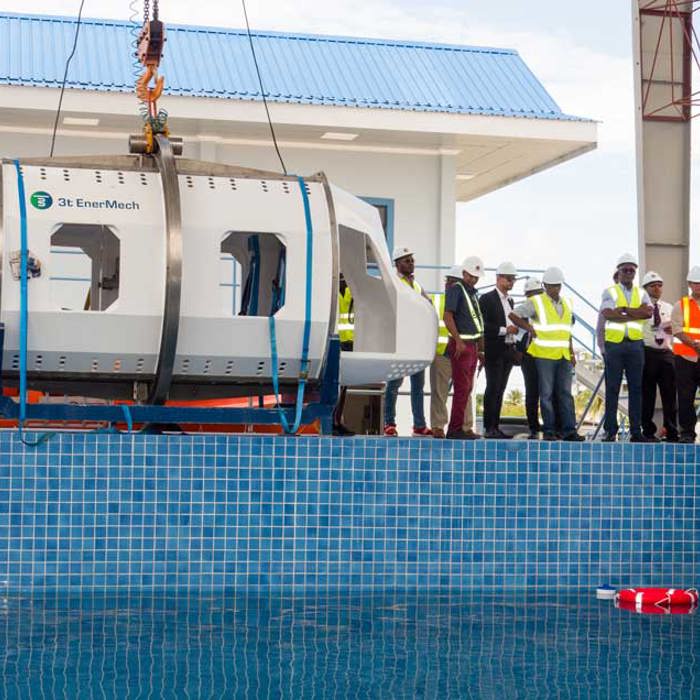 A group of workers in safety gear observe a helicopter evacuation simulator being lowered into a pool.