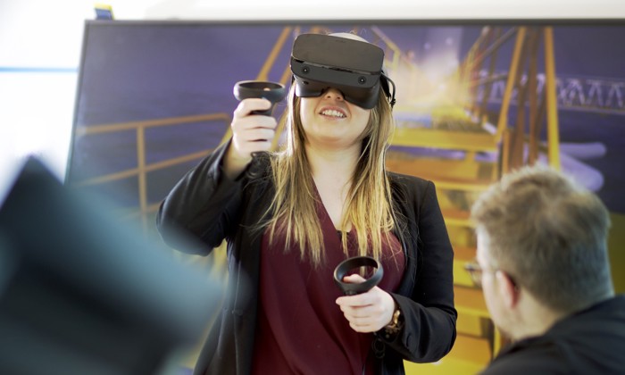 A woman wearing a virtual reality headset and holding controllers interacts with a digital simulation, while a man observes in the foreground.