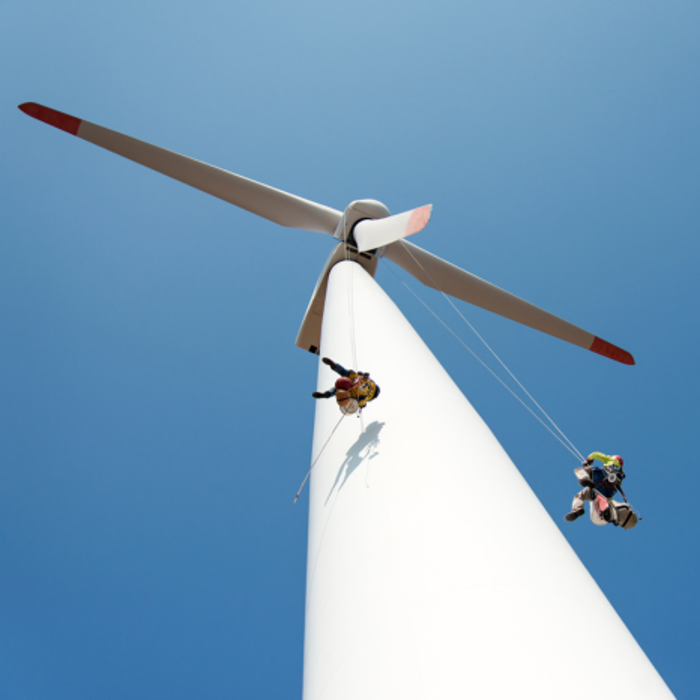 Two technicians, well-trained and suspended by ropes, work diligently on a large wind turbine against the backdrop of a clear blue sky.