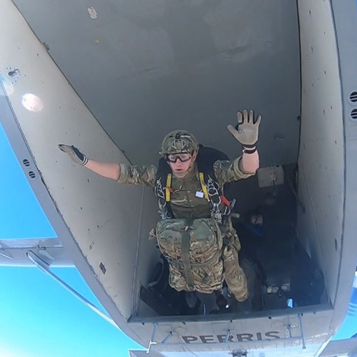 A soldier in full gear, including a parachute, exits from the rear of an aircraft during a skydive, seen waving with one hand, with a clear blue sky in the background.