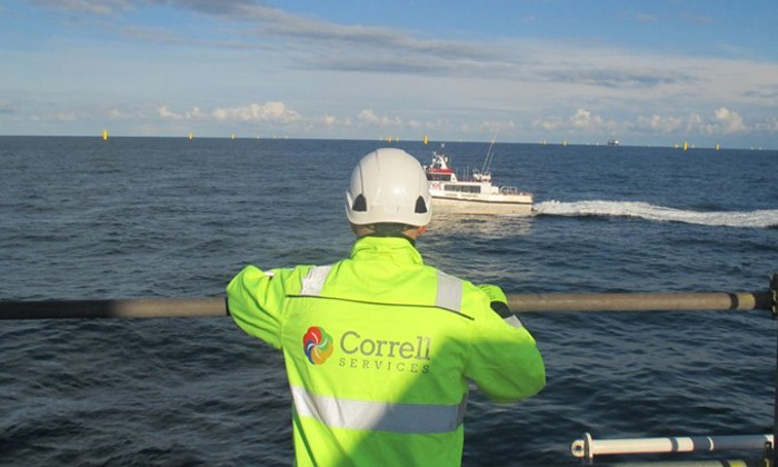 A person in a high-visibility jacket and helmet watches a boat in the ocean. The jacket has a "Correll Services" logo on the back.