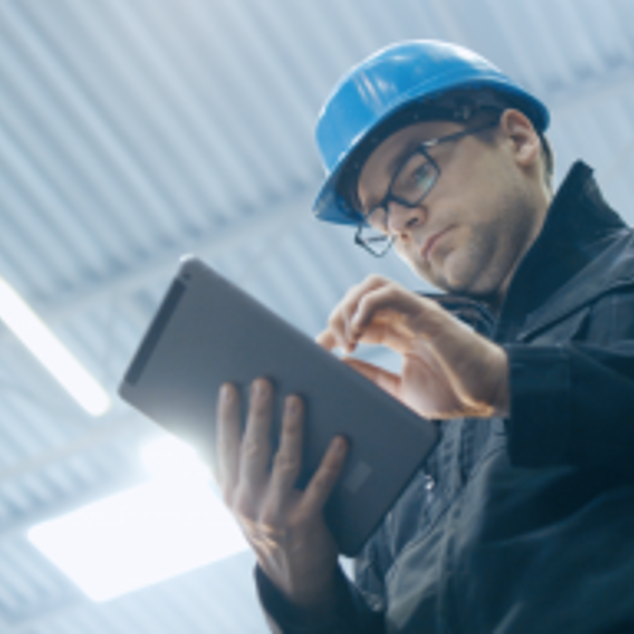 A person wearing a hard hat and glasses uses a tablet in an industrial setting with a corrugated metal ceiling.