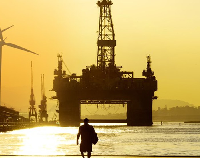 Silhouette of a person walking towards a large offshore oil rig at sunset, with wind turbines visible in the background.