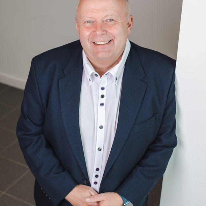 Bald man in a navy suit and white shirt, smiling, standing indoors against a light-colored wall.