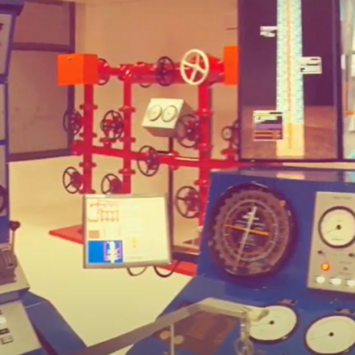 A control room with various dials, gauges, and equipment panels, situated behind a valve system mounted on a red frame.