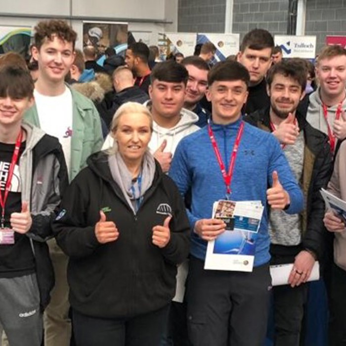A group of people, mostly young men with a woman in the center, give thumbs up at an indoor event with informational booths and crowds in the background. They have event badges and brochures.
