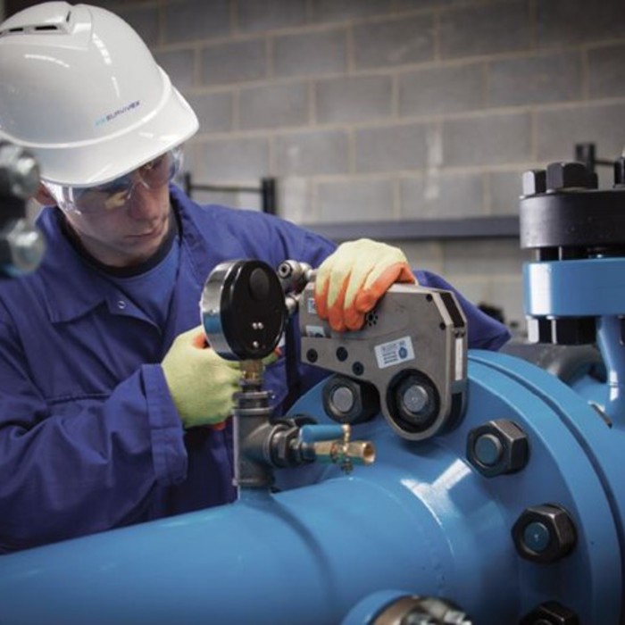 A worker in a blue jumpsuit and white helmet uses equipment to perform maintenance on blue industrial piping in a facility with a concrete block wall.