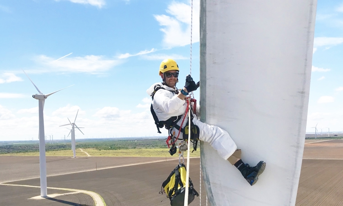 A worker in safety gear, including a helmet and harness, is suspended on a rope while performing maintenance on a large wind turbine blade with several wind turbines in the background, highlighting the military to wind transition in skilled labor.