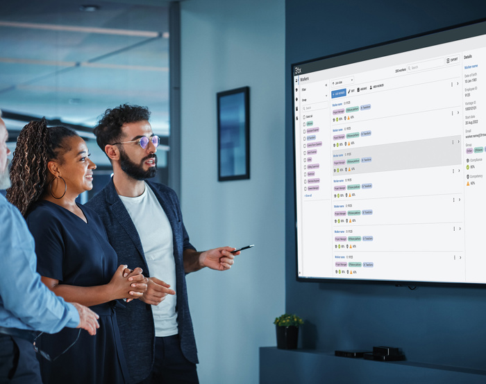 Three colleagues stand in front of a large screen displaying project management software, discussing the details and pointing at the screen.