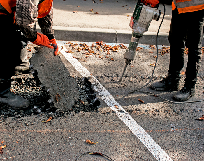 Two workers in safety gear, adept from rigorous training, break and remove a section of asphalt on a road using a jackhammer.