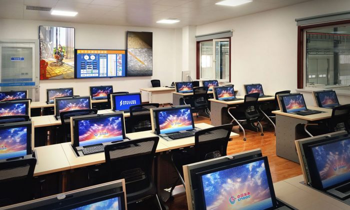 A modern classroom equipped with numerous workstations, each featuring a computer displaying a colorful screensaver. A large digital screen and whiteboard are visible at the front of the room.
