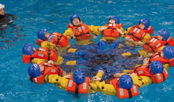 A group of people in life jackets and helmets form a circle in a pool as part of a training session. A person in black observes from the side.