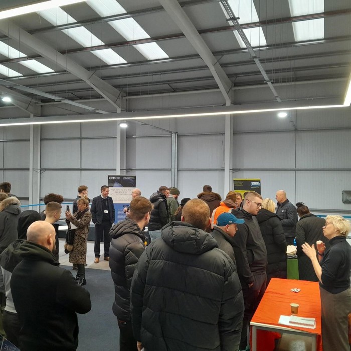 A group of people gathered indoors for a training event, near display tables and banners. Many are wearing jackets. The space has high ceilings and overhead lighting, creating an inviting atmosphere for learning and networking.