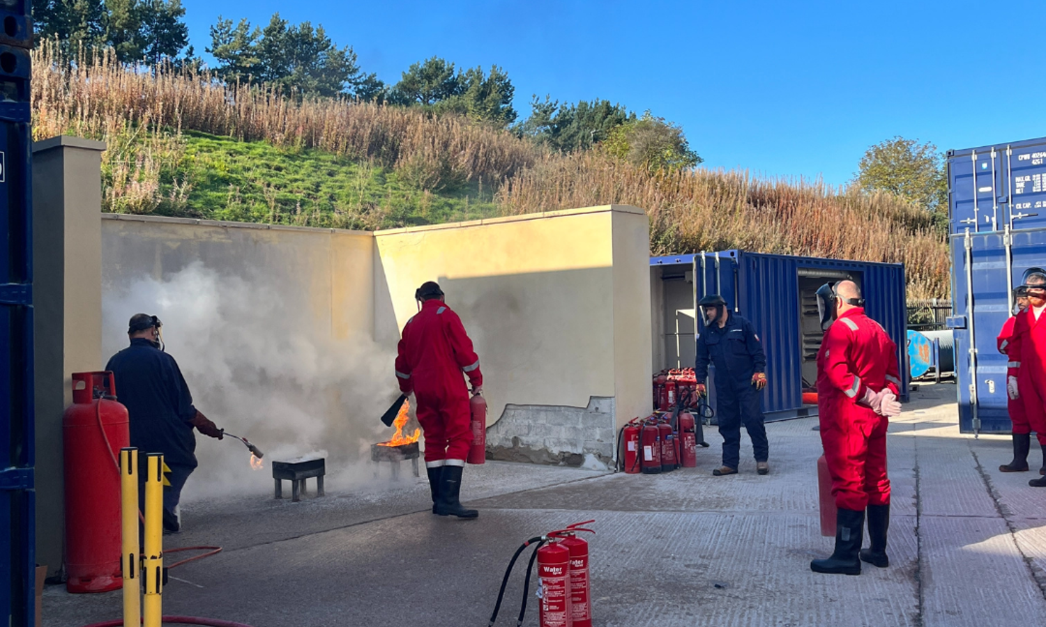 In a dedicated training zone, people in red uniforms practice extinguishing a fire outdoors, surrounded by visible fire extinguishers and safety equipment.