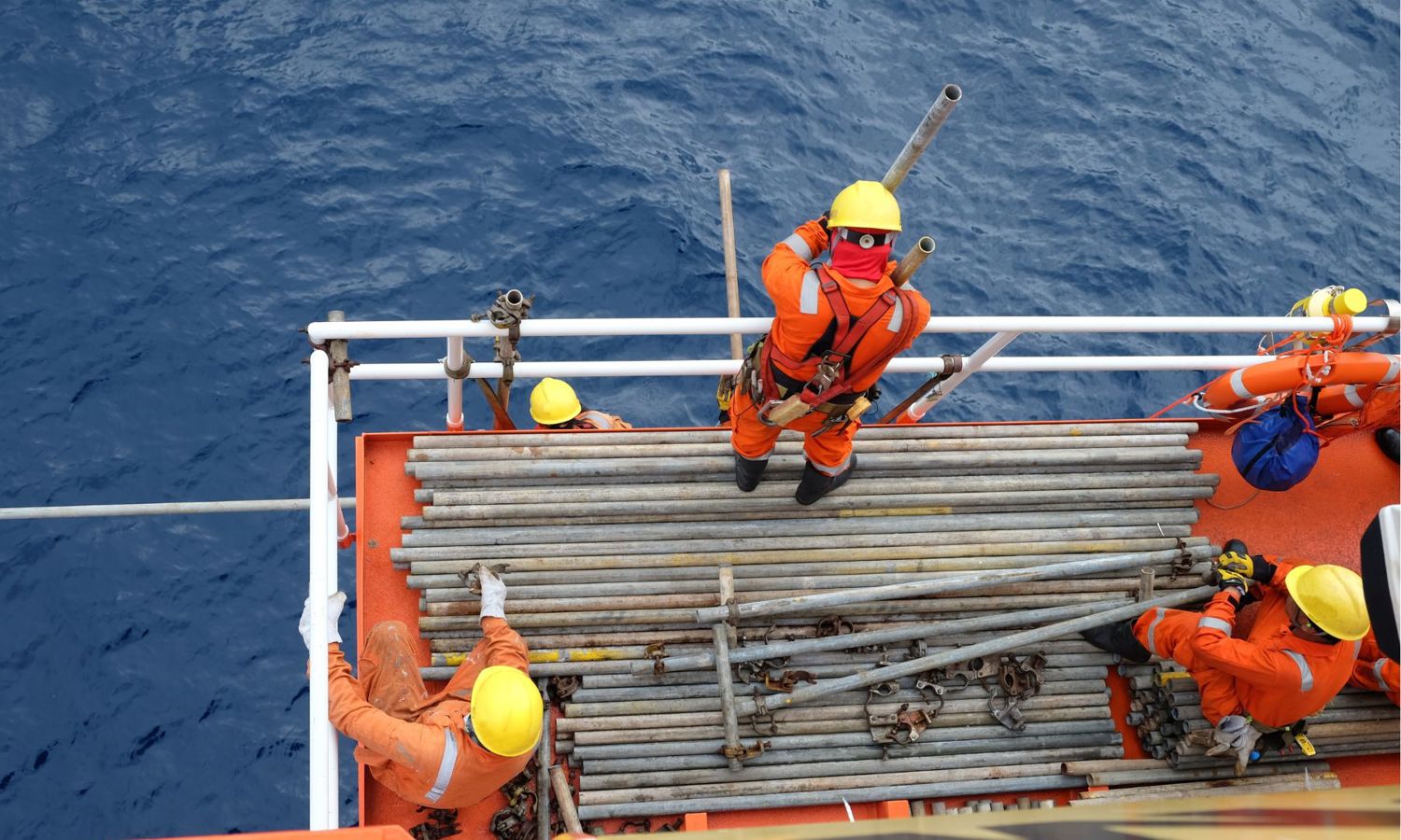 Workers in orange safety gear and yellow helmets handle metal pipes on the deck of an offshore platform against a backdrop of the ocean.